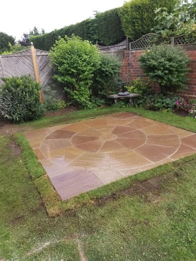 Patio area with stone slabs surrounded by grass and shrubs in a garden setting.