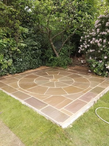 A circular patterned stone patio surrounded by greenery and flowering plants.