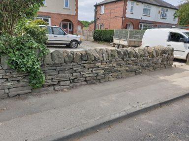 Stone wall alongside a pavement, with cars parked nearby and houses in the background.