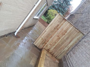 A narrow path beside a house leads to a wooden fence on a rainy day.