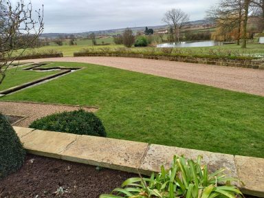 Lush green lawn with a gravel path, bordered by shrubs and distant trees.