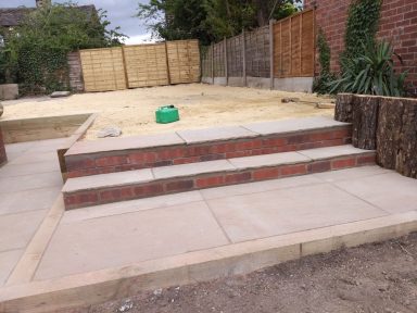 Brick steps lead to a paved area with a sandy surface and wooden fencing.