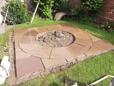 A circular stone patio partially completed in a garden, surrounded by grass and plants.