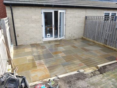 Paved outdoor area with sandstone slabs, adjacent to a house and wooden fence.